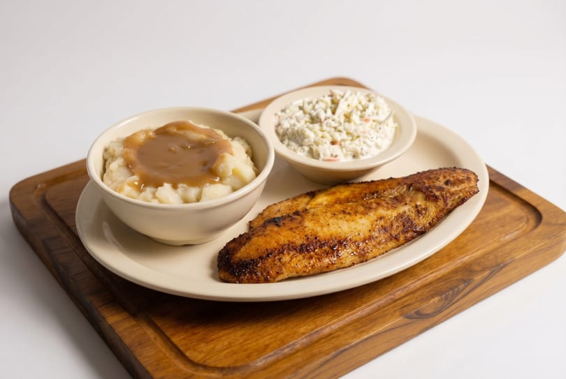 A plate of grilled fish, mashed potatoes with gravy, and coleslaw on a wooden board.