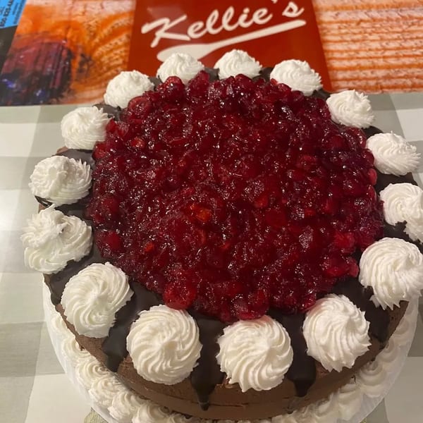 A chocolate cake with cherry topping and whipped cream dollops, with a "Kellie's" sign in the background.