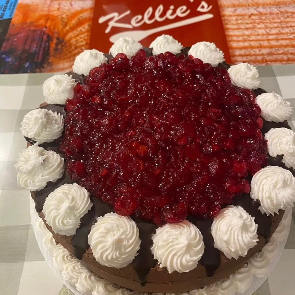 A chocolate cake with cherry topping and whipped cream dollops, with a "Kellie's" sign in the background.