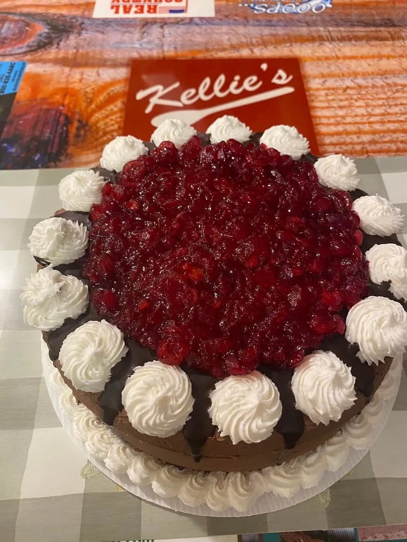 A chocolate cake with cherry topping and whipped cream dollops, with a "Kellie's" sign in the background.